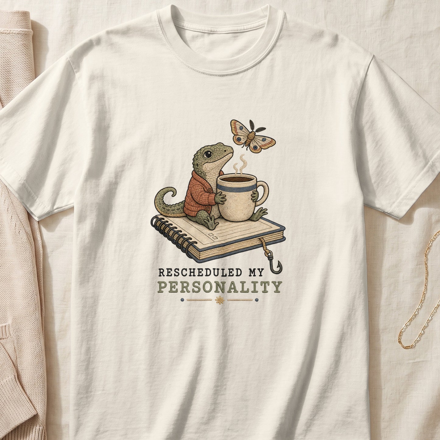 Off-white t-shirt featuring an illustration of a lizard wearing a sweater, holding a mug, sitting on a notebook, with a moth and the text Rescheduled My Personality.