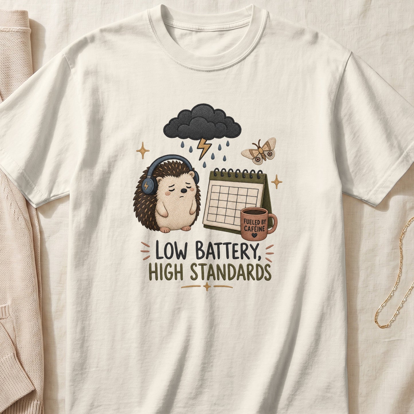 T-shirt graphic with a sad hedgehog, calendar, coffee cup, and the text "LOW BATTERY, HIGH STANDARDS".