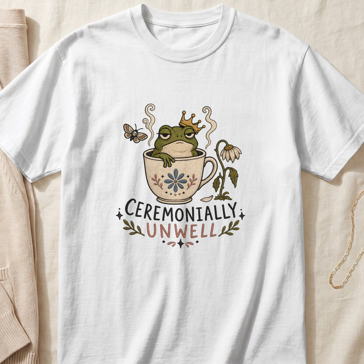 White t-shirt with a graphic of a crowned frog in a teacup, a moth, and the text "CEREMONIALY UNWELL".