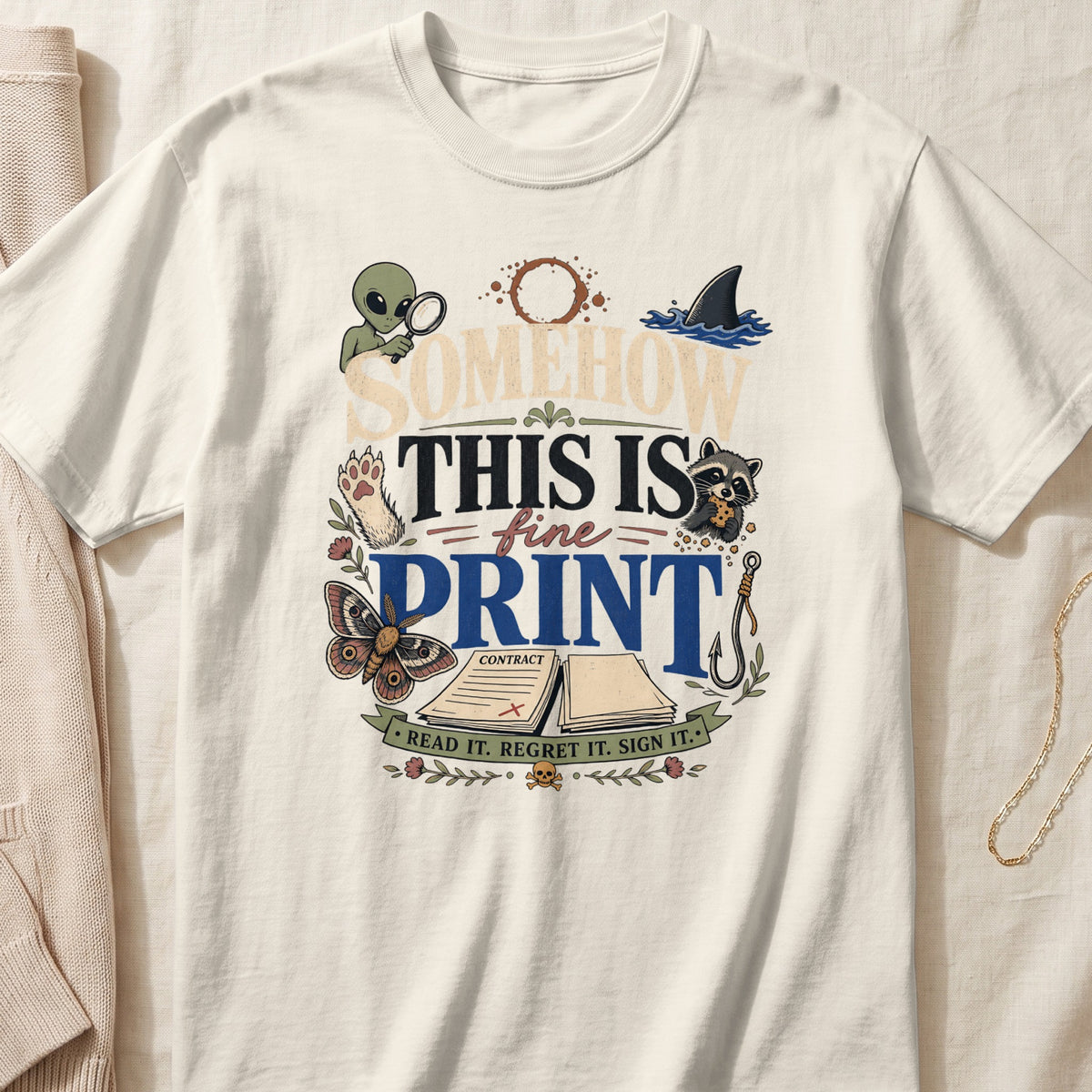 A cream-colored t-shirt with a vintage graphic containing the text "SOMEHOW THIS IS FINE PRINT" and various whimsical illustrations.