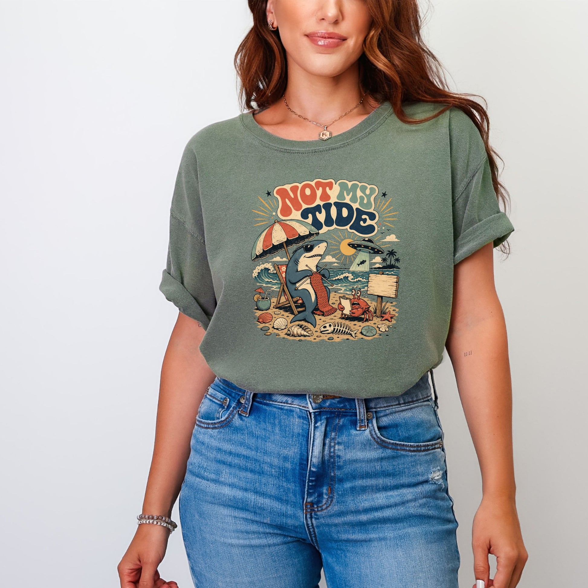 T-shirt graphic: Shark knitting on a beach with text "NOT MY TIDE" and a UFO.