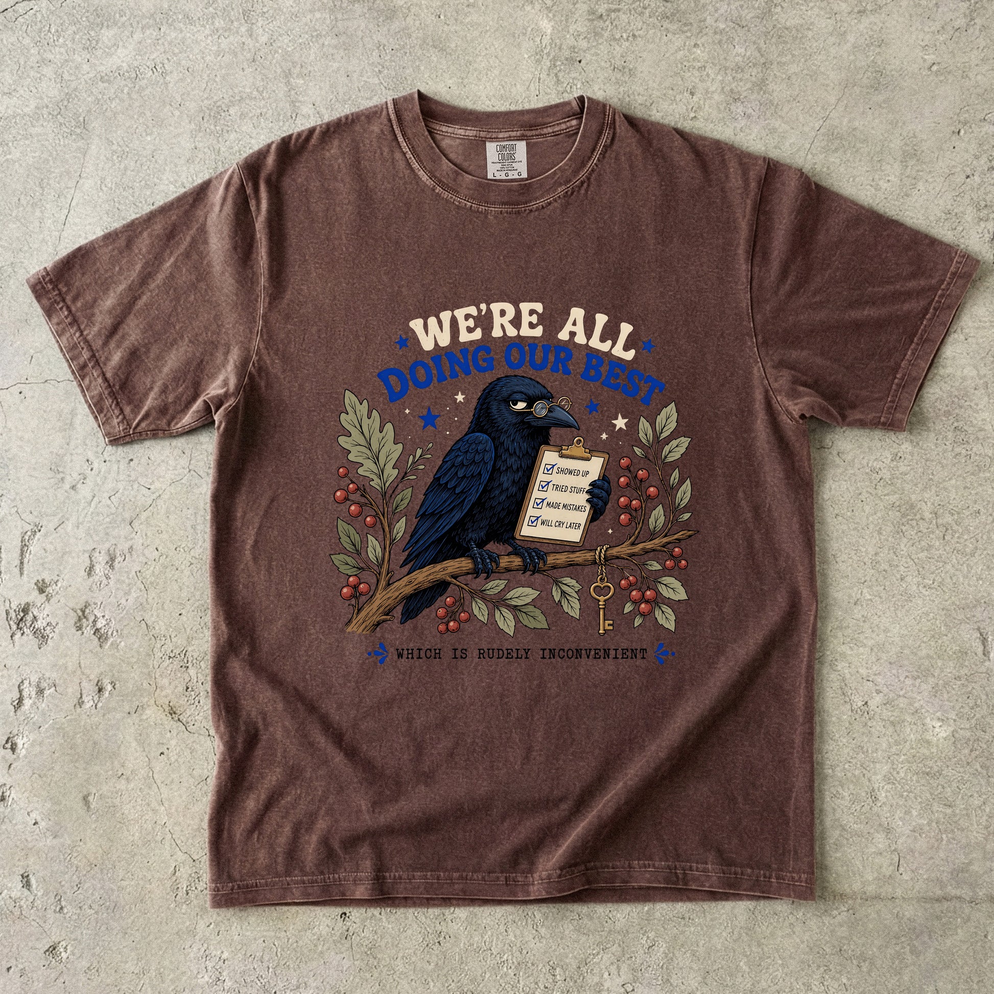 Brown t-shirt with a crow holding a clipboard that says "We're all doing our best".
