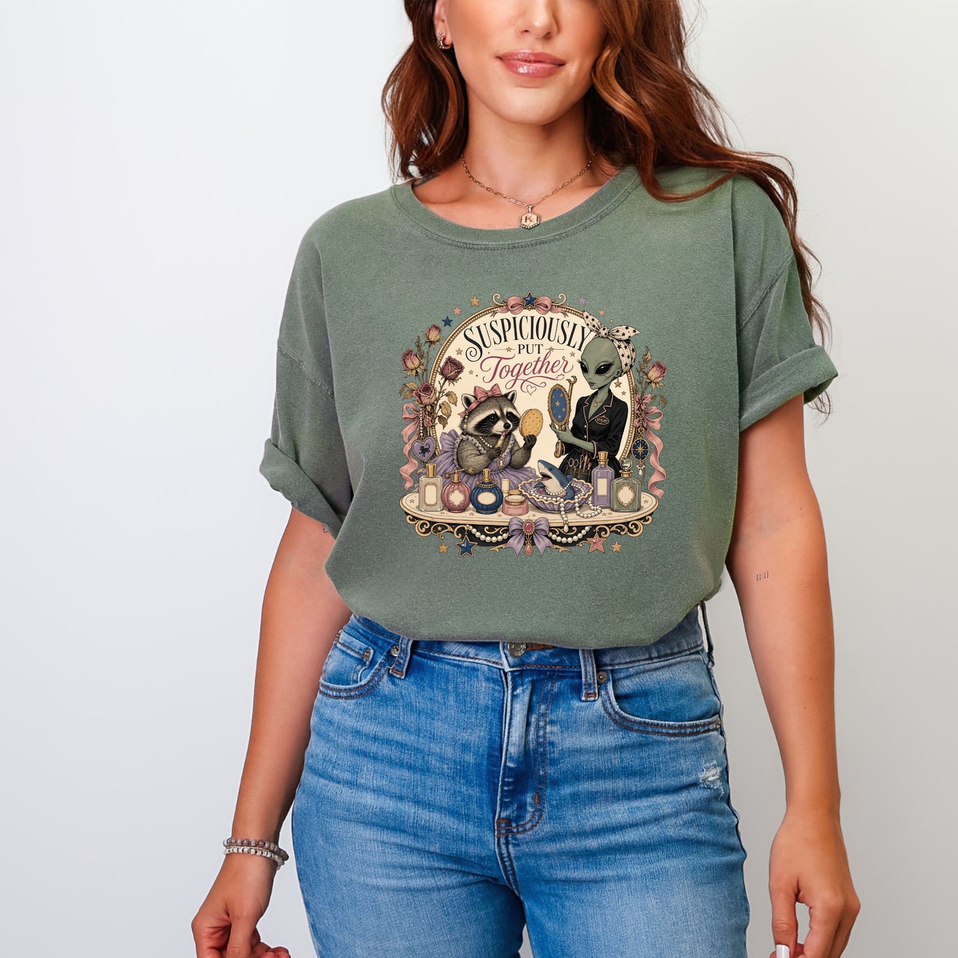 A woman wearing an olive-green t-shirt printed with an illustration of an alien and a raccoon and the text "Suspiciously Put Together".