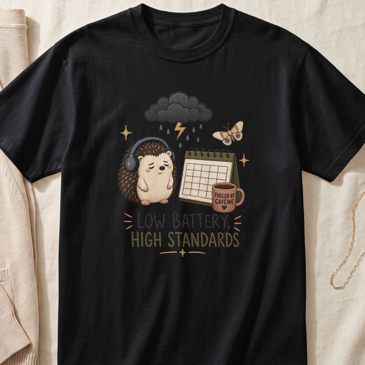 A black t-shirt features a graphic of a tired hedgehog wearing headphones beneath a rain cloud with text reading Low Battery High Standards.