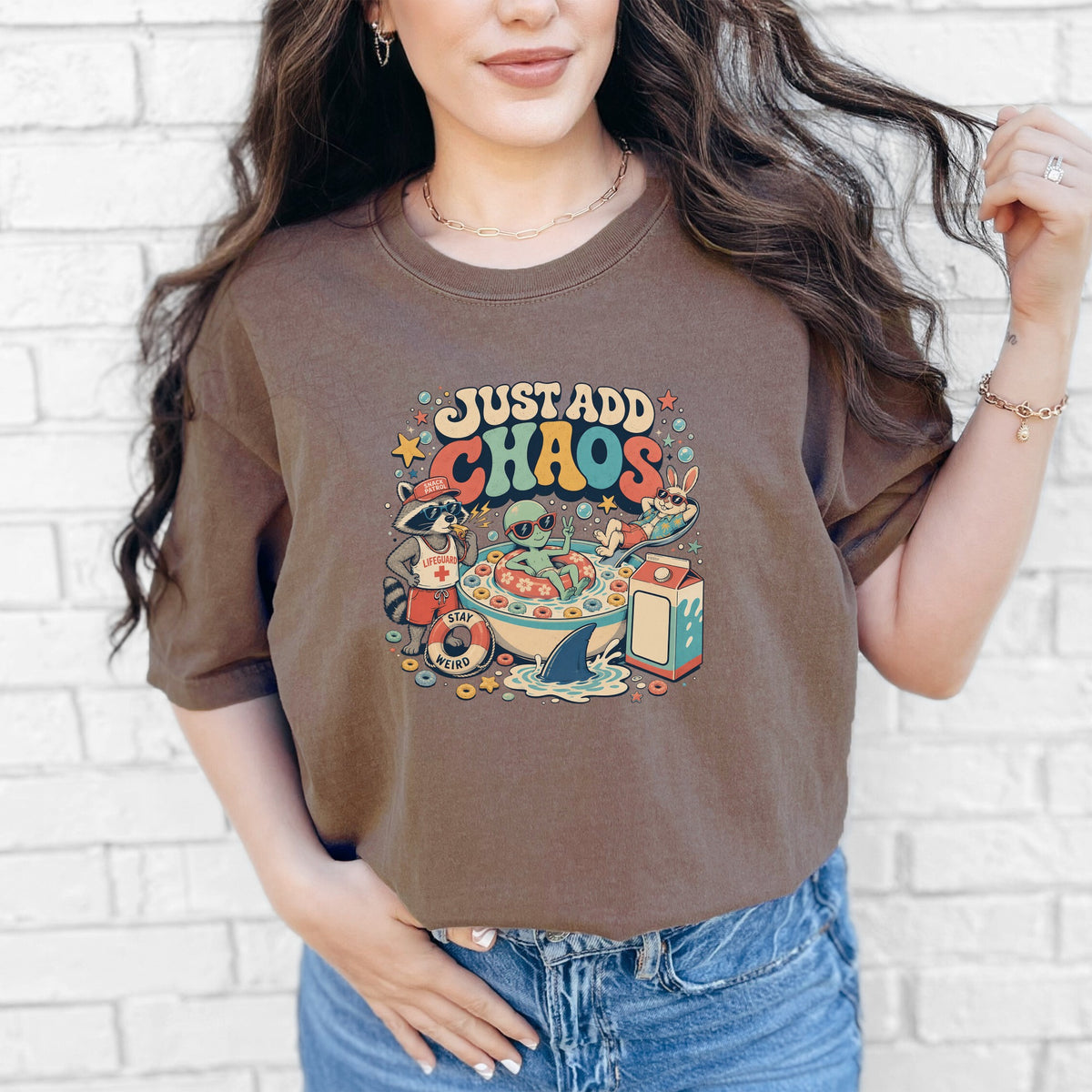 A woman wears a brown t-shirt featuring a cartoon graphic of an alien in a cereal bowl with the text Just Add Chaos.