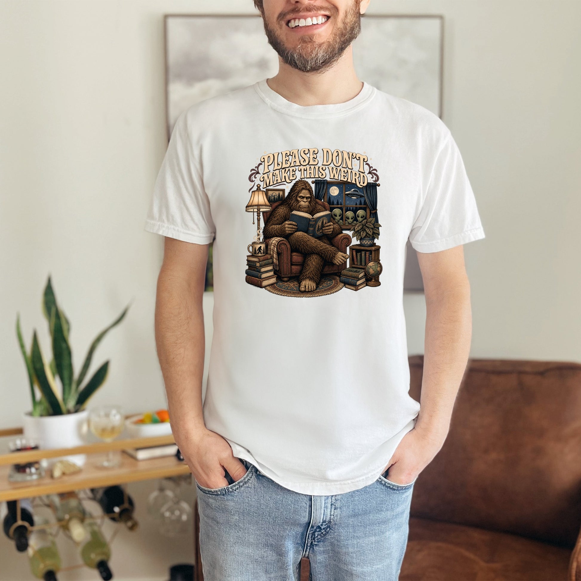 White t-shirt graphic with Bigfoot reading surrounded by aliens and text "Please Don't Make This Weird".