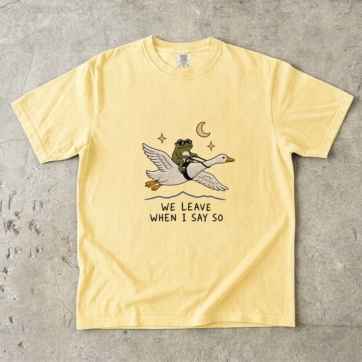 Yellow t-shirt with graphic of frog riding a goose under moon and stars, text "WE LEAVE WHEN I SAY SO".
