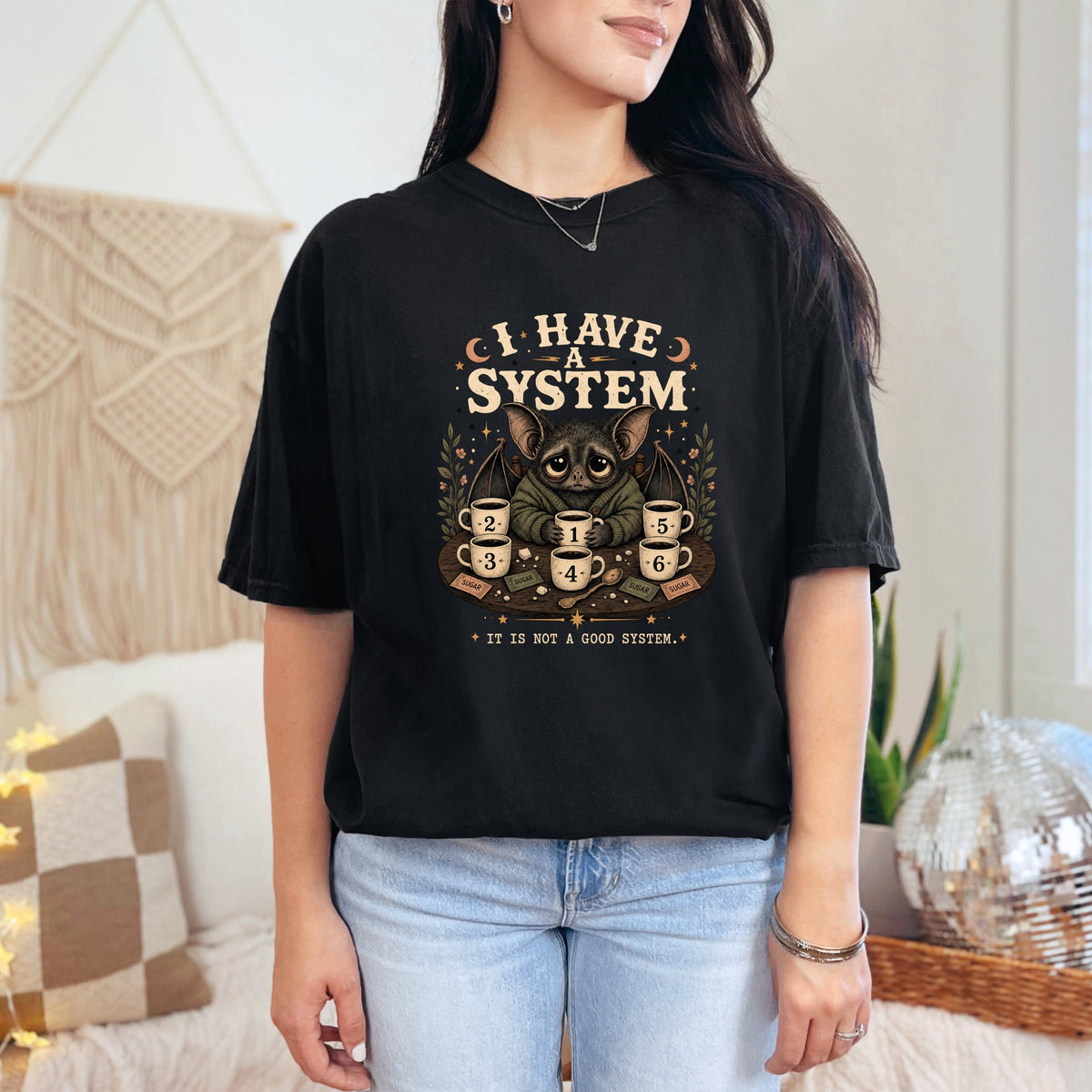 Black t-shirt graphic with bat holding coffee mugs numbered 1-6 and text "I HAVE A SYSTEM. IT IS NOT A GOOD SYSTEM."
