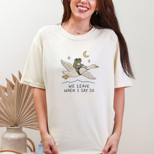 Cream t-shirt with a graphic of a frog riding a flying goose, with stars, moon, and text.