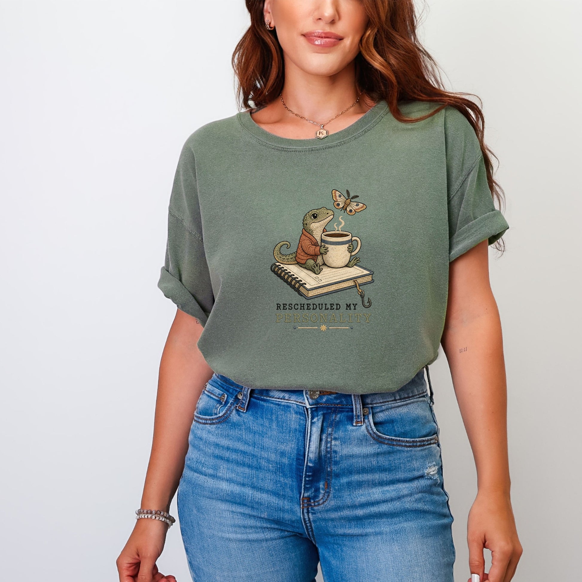 A woman wearing an olive-green t-shirt printed with a graphic of a lizard holding a coffee mug on a notebook, with text reading "RESCHEDULED MY PERSONALITY."
