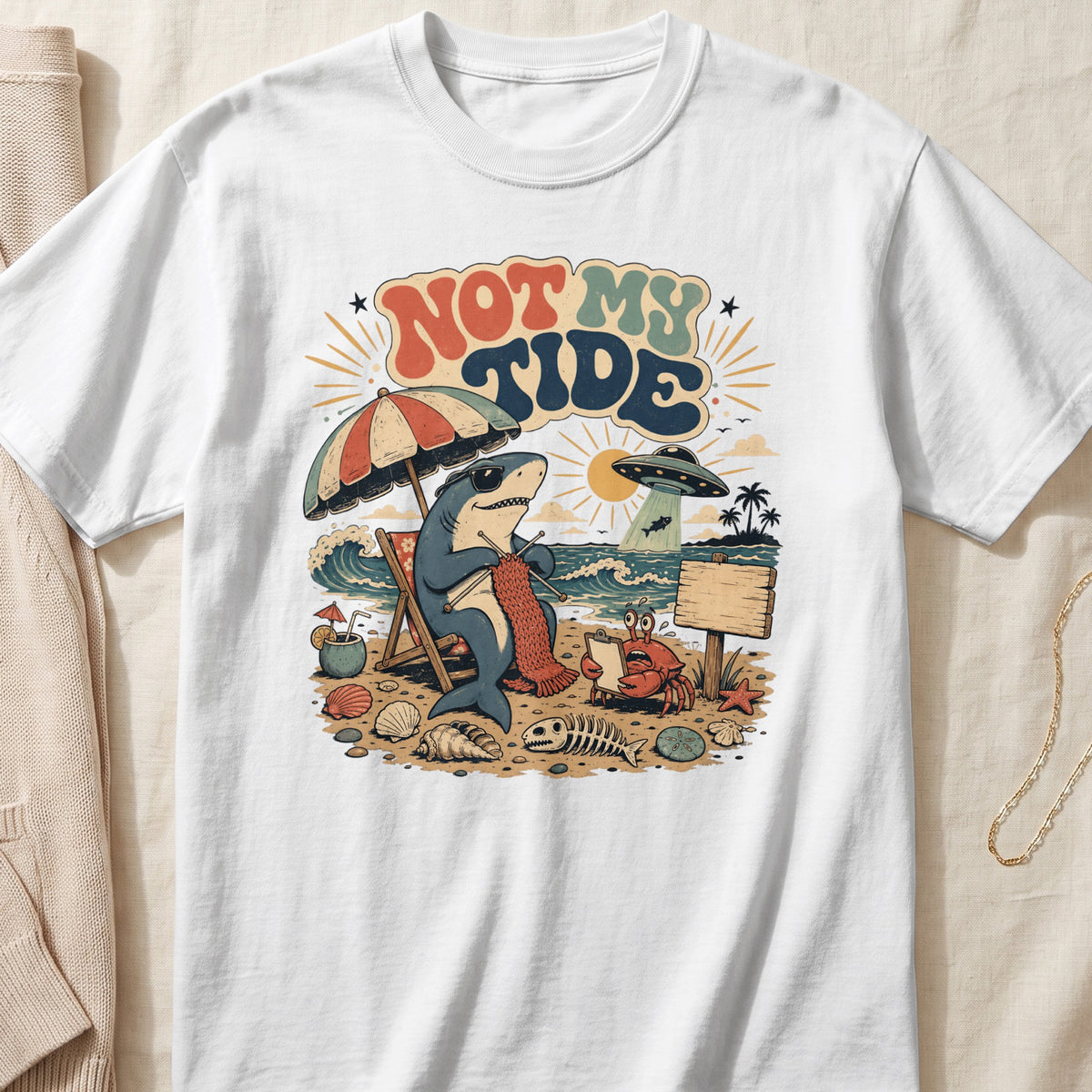 Shark knitting on a beach chair with "Not My Tide" text, UFO, and crab.