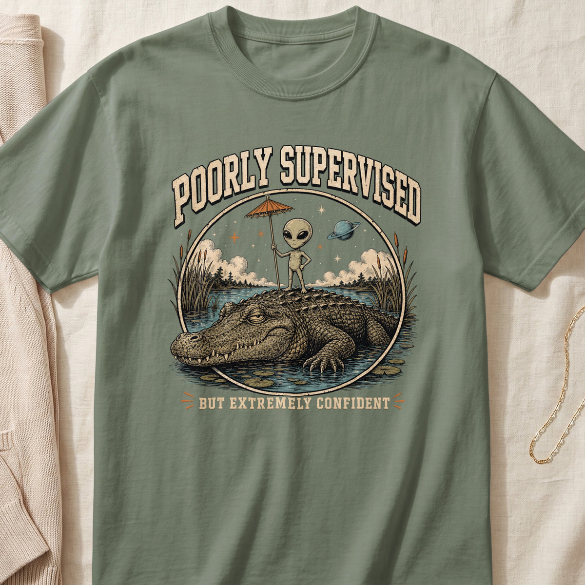 Green t-shirt with alien on alligator, holding umbrella, "Poorly Supervised But Extremely Confident."