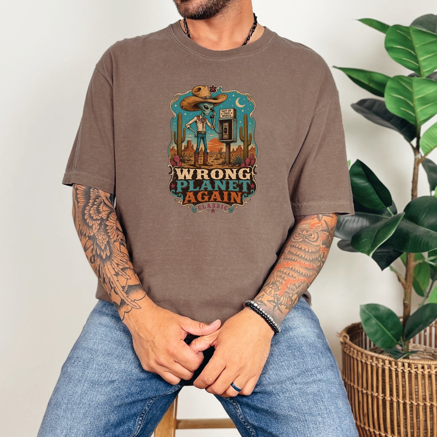 A tattooed man wearing a brown t-shirt featuring a vintage alien cowboy graphic and blue jeans.