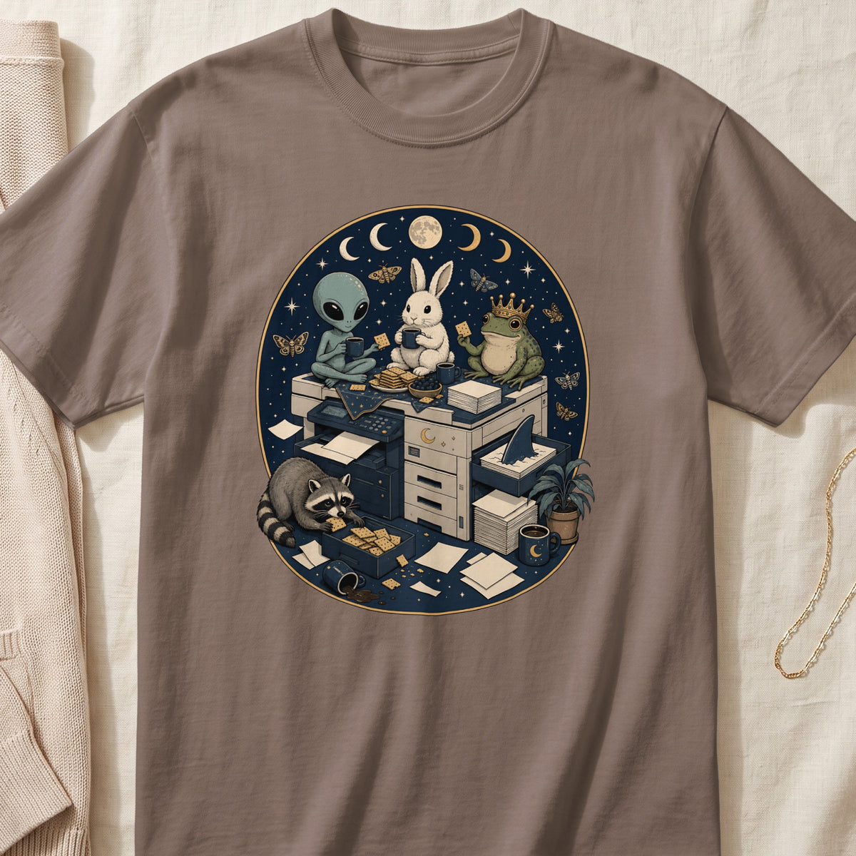 Brown t-shirt featuring a circular celestial illustration of an alien, rabbit, frog, and raccoon gathered around an office printer.