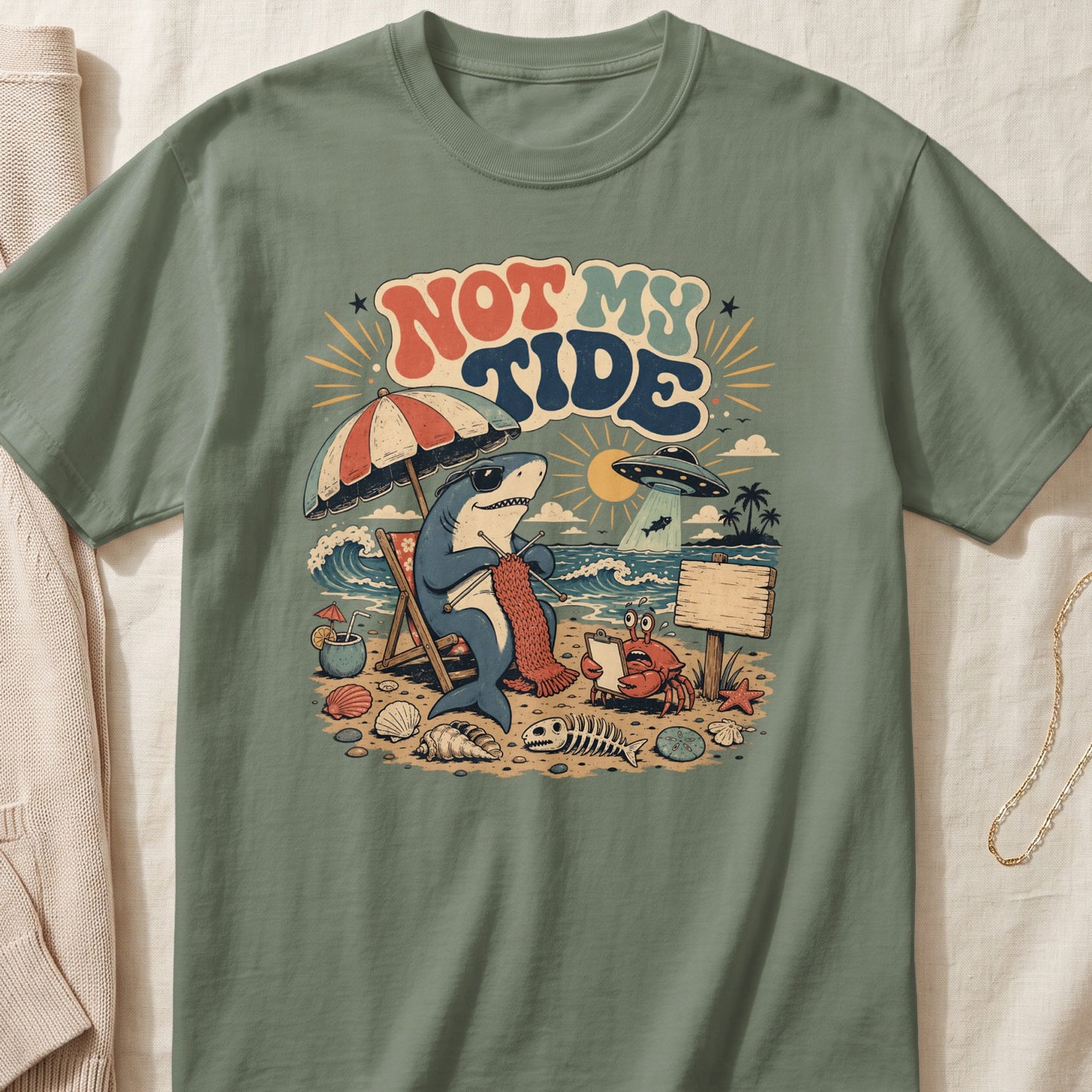 Shark knitting on beach, with crab, UFO, and text "NOT MY TIDE."