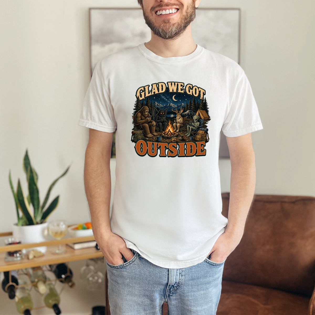 White t-shirt with Bigfoot, mothman, alien, and deer around campfire text: Glad we got outside.