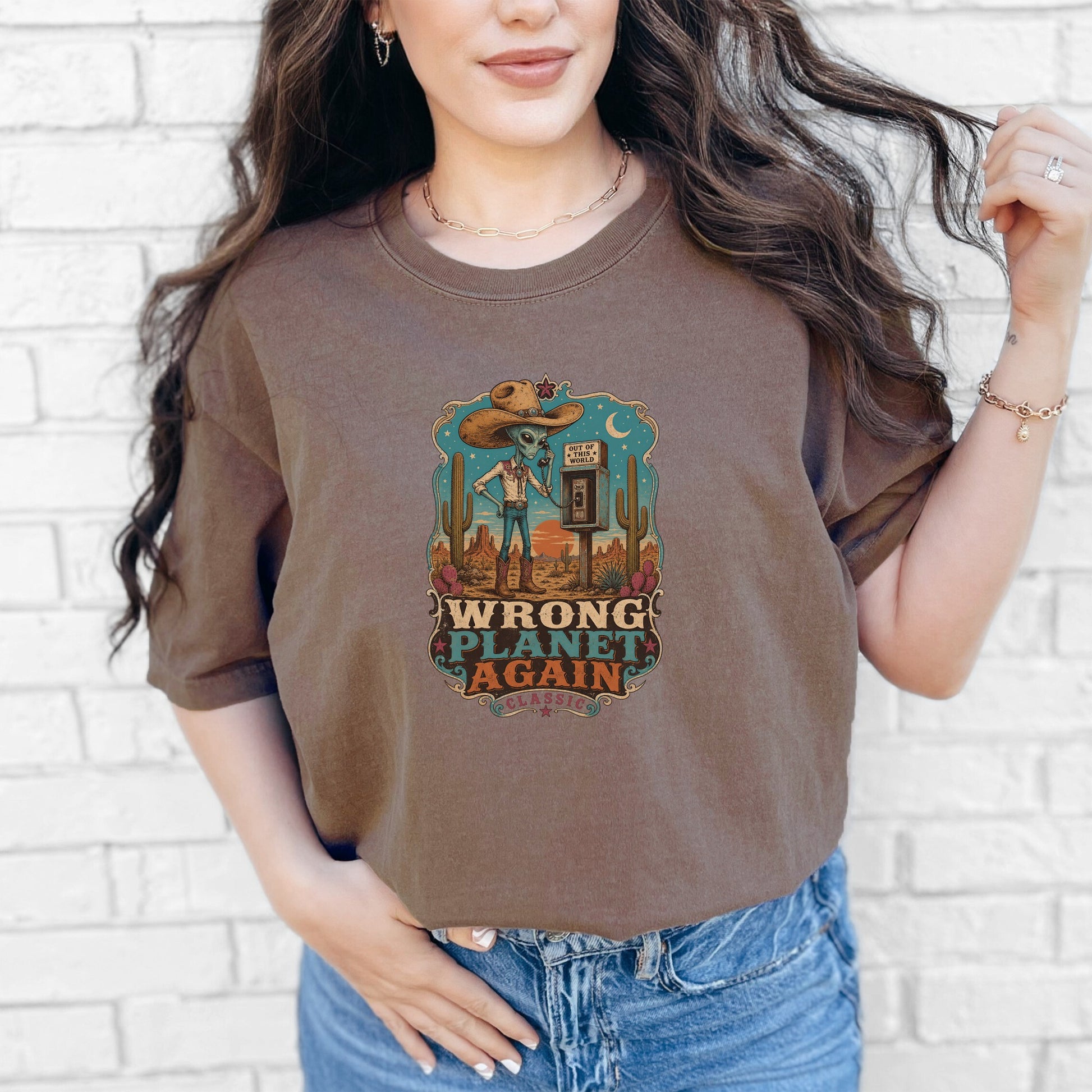 A woman wears a brown t-shirt featuring a vintage western graphic that reads Wrong Planet Again.