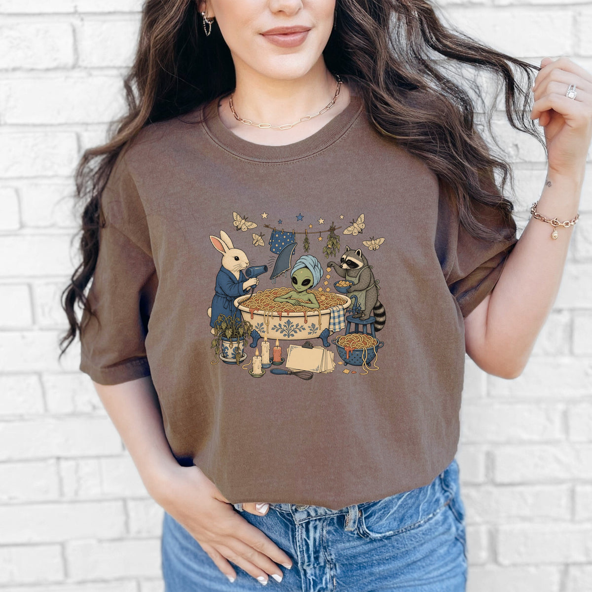 A woman wearing a brown t-shirt featuring an illustrated alien relaxing in a spaghetti bath attended by a rabbit and raccoon.