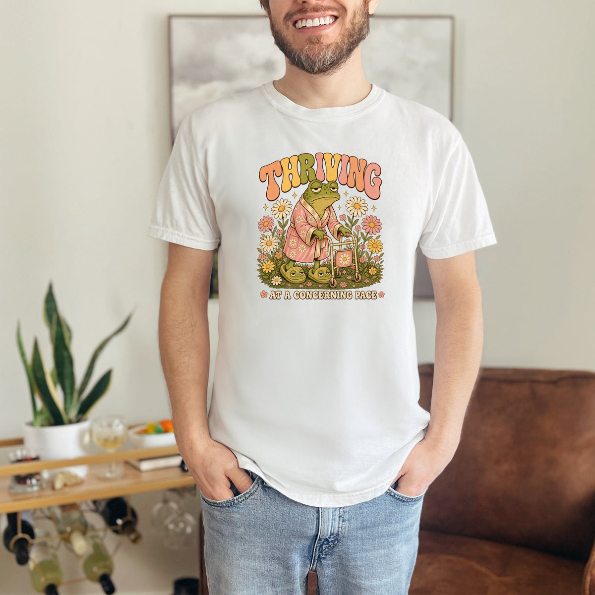 White t-shirt with graphic of frog in robe, walker, and text "THRIVING AT A CONCERNING PACE".