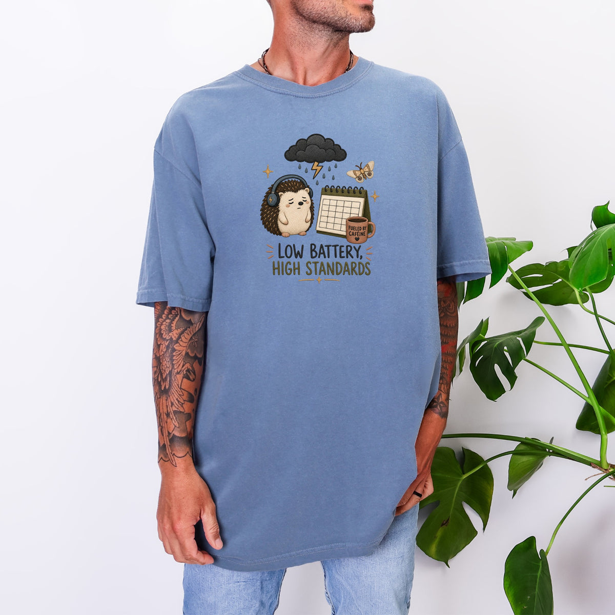 A man wearing an oversized blue t-shirt featuring an embroidered graphic of a sleepy hedgehog, calendar, and text that reads "LOW BATTERY, HIGH STANDARDS".