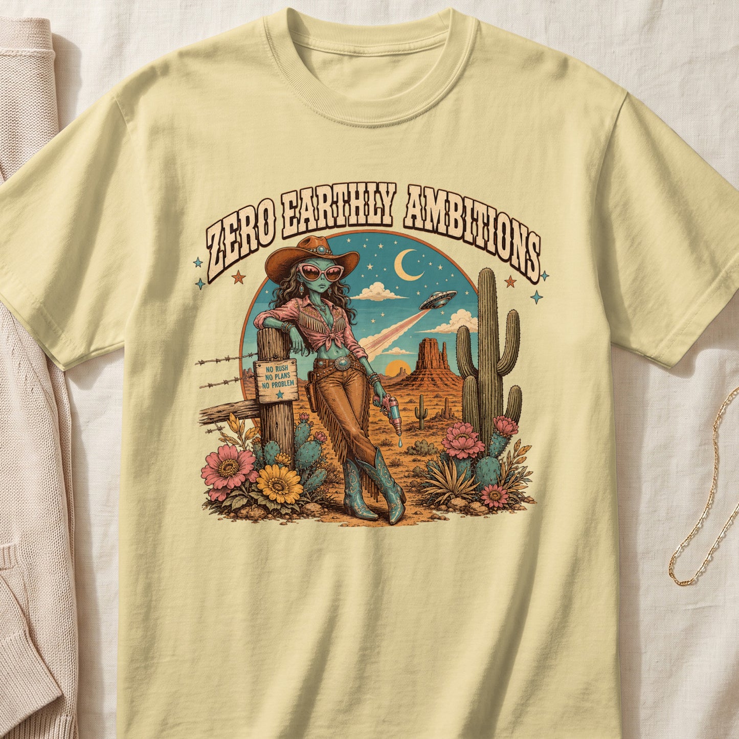 T-shirt graphic with alien cowgirl in desert, text "ZERO EARTHLY AMBITIONS".