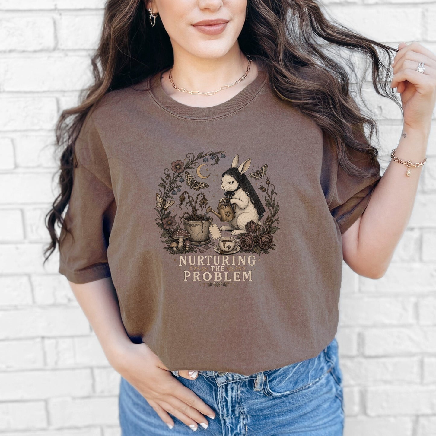 A woman wears a brown crewneck t-shirt featuring a rabbit watering a plant and the text NURTURING THE PROBLEM.