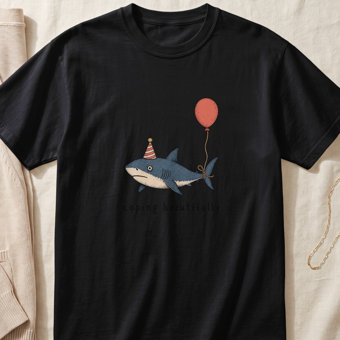 Black t-shirt with a drawing of a shark wearing a party hat and holding a balloon with text "coping beautifully".