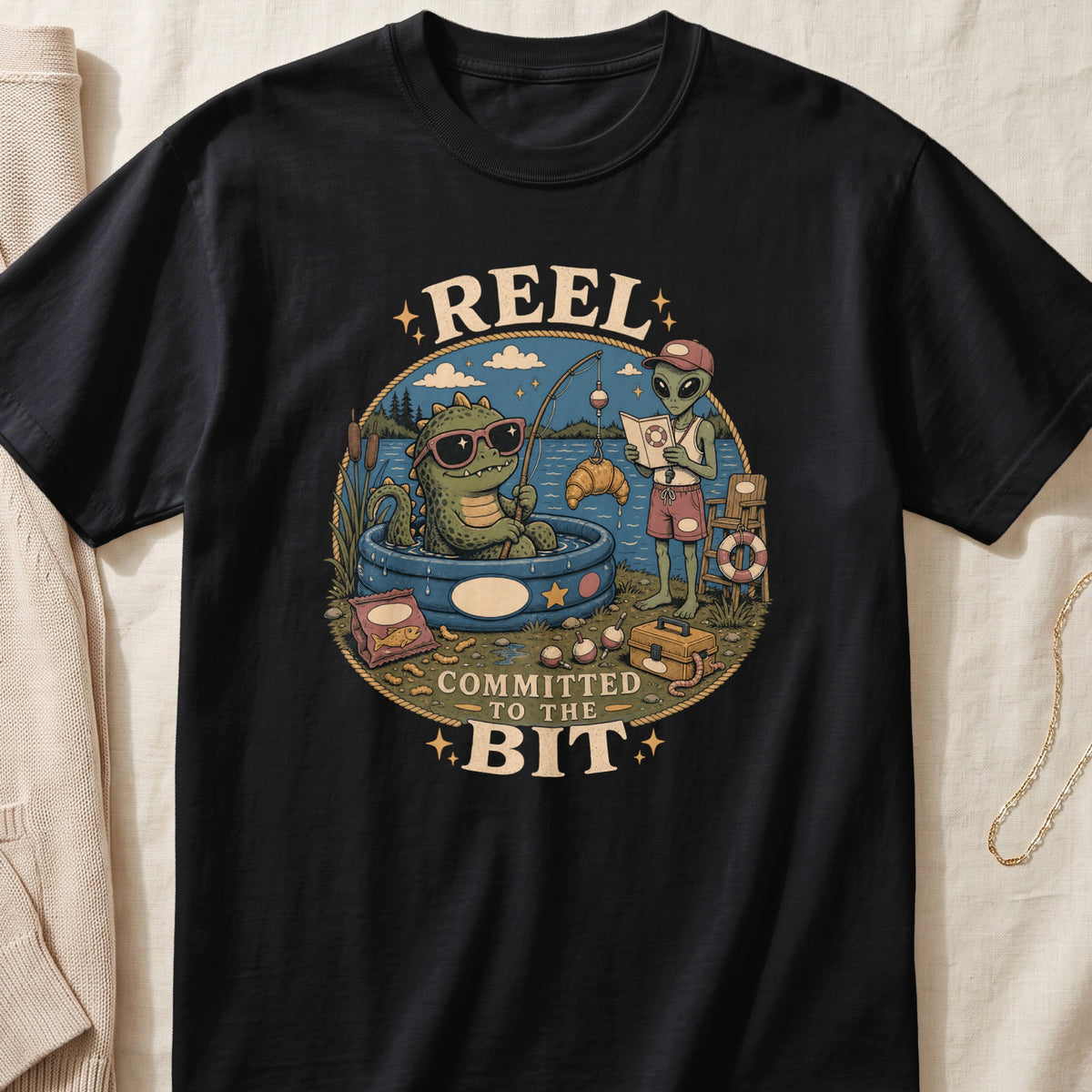 Black t-shirt featuring cartoon dragon and alien fishing with text "REEL COMMITTED TO THE BIT".