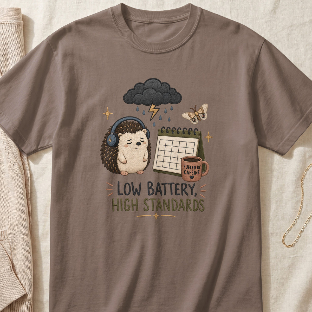 Brown t-shirt with embroidered sad hedgehog, storm cloud, calendar, and "Low Battery, High Standards" text.