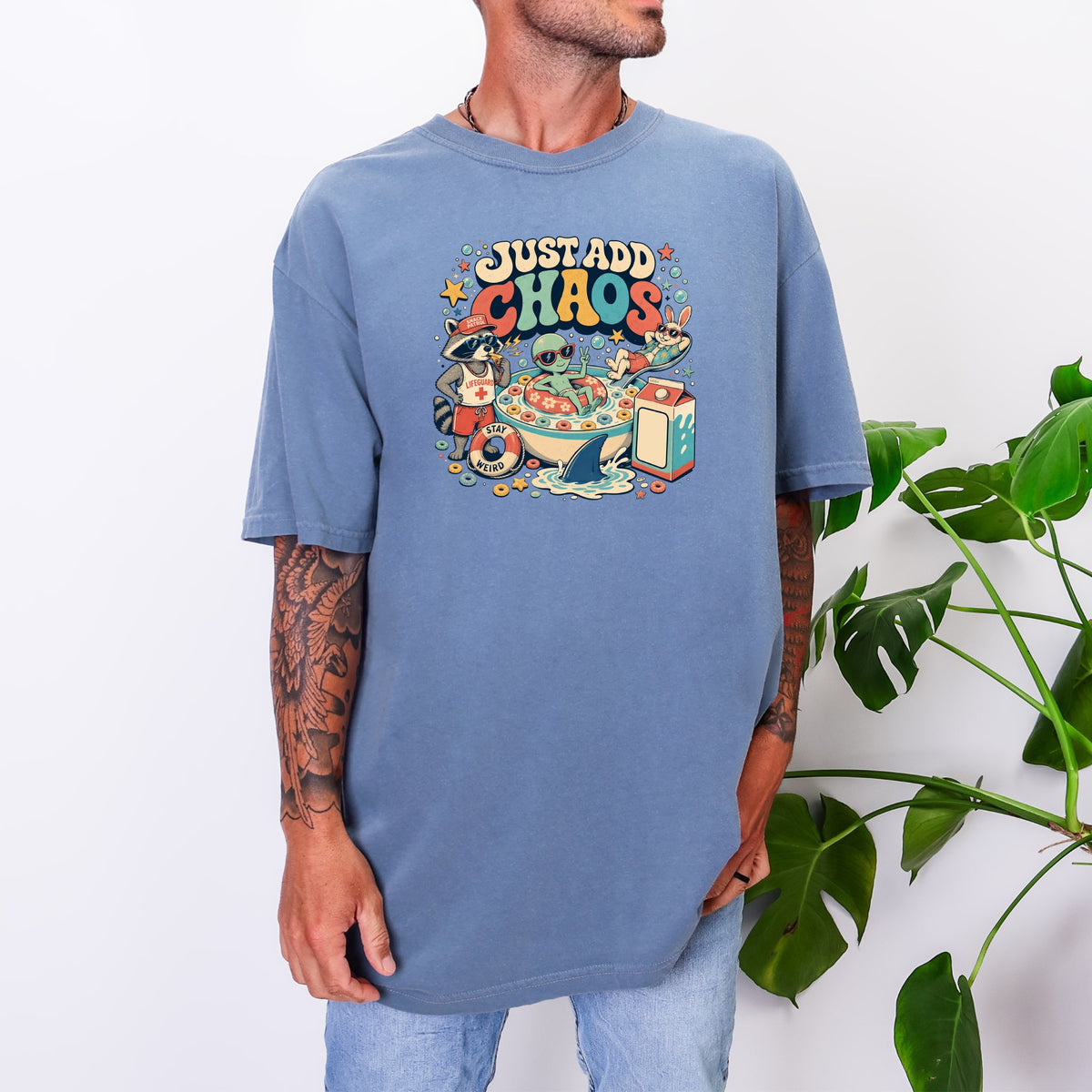 A man wears an oversized blue t-shirt featuring a colorful graphic of cereal and cartoon animals with the text "Just Add Chaos."