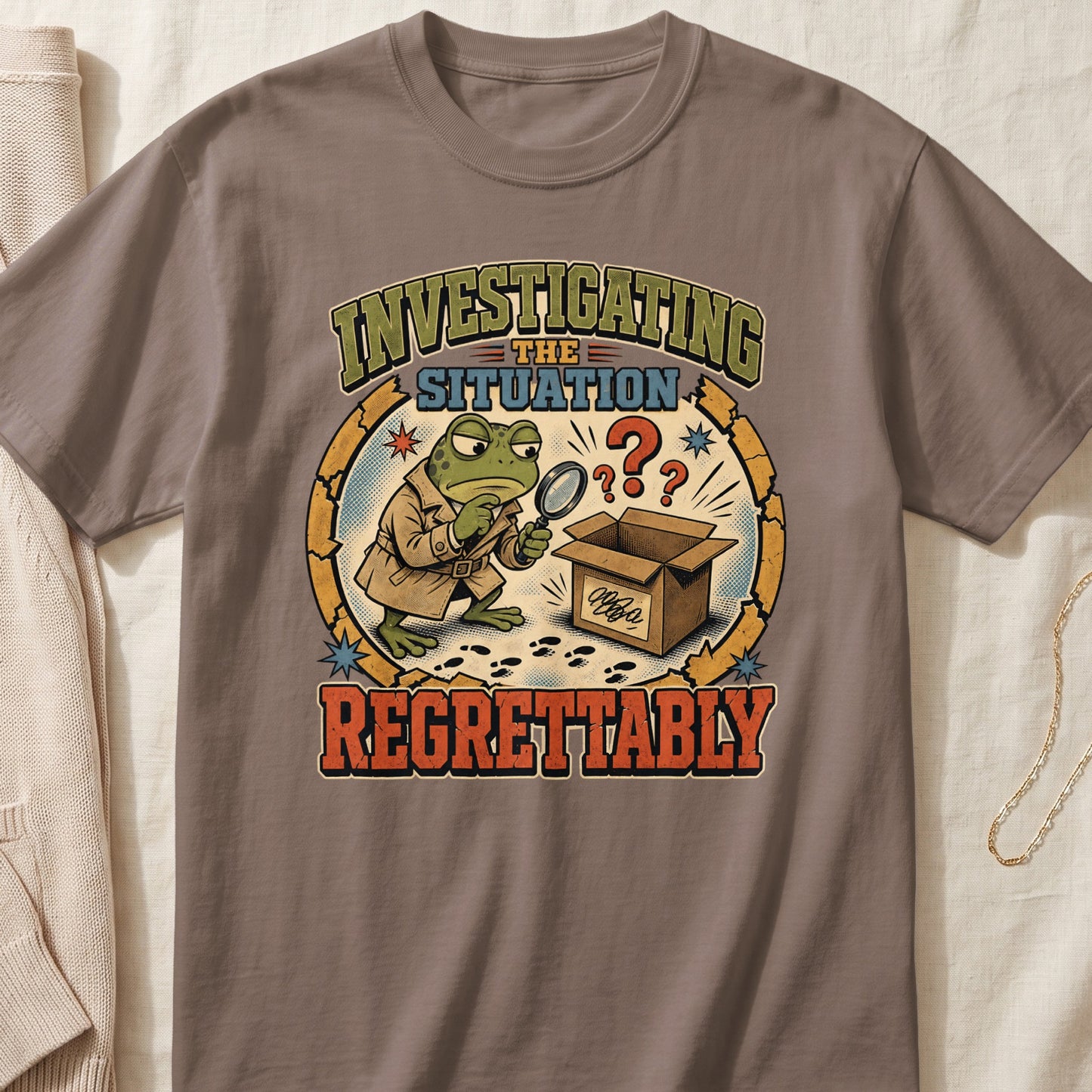 Brown t-shirt featuring a cartoon frog detective examining an open box with a magnifying glass.
