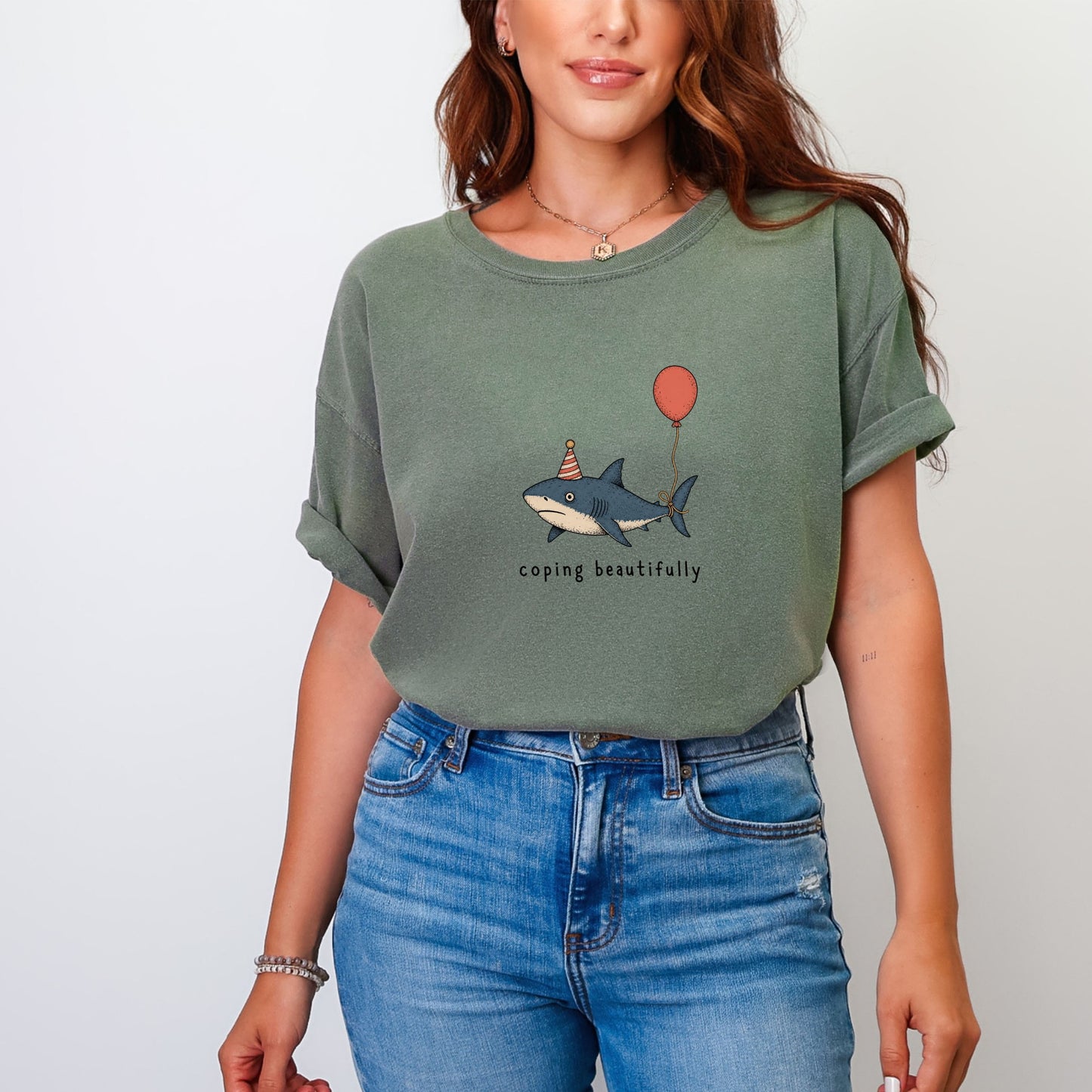 Green t-shirt with a shark wearing a party hat and holding a balloon with the text "coping beautifully".