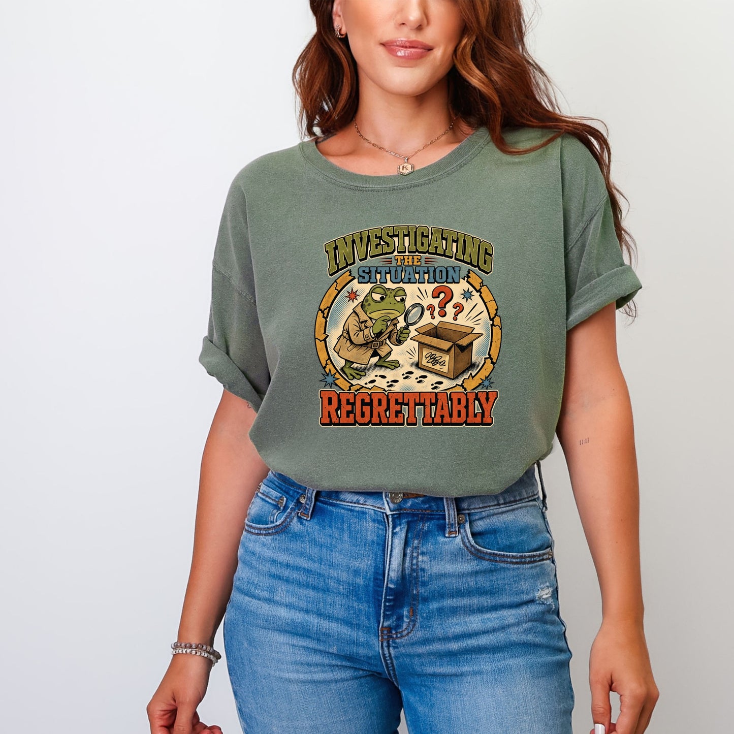 A person wearing an olive green t-shirt featuring a cartoon frog looking into a cardboard box with a magnifying glass.