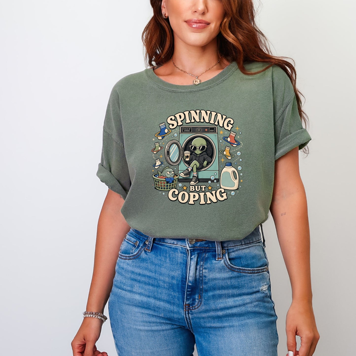 A woman wearing an olive-green t-shirt with a graphic of an alien sitting inside a washing machine, surrounded by text that reads "Spinning But Coping."