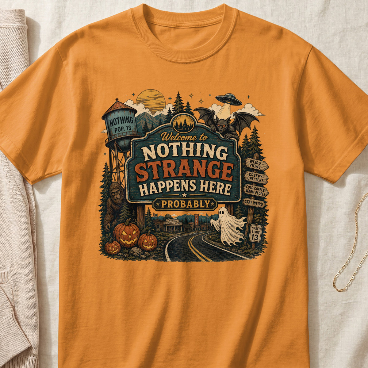 Orange t-shirt featuring "Welcome to Nothing Strange Happens Here Probably" sign with spooky elements.