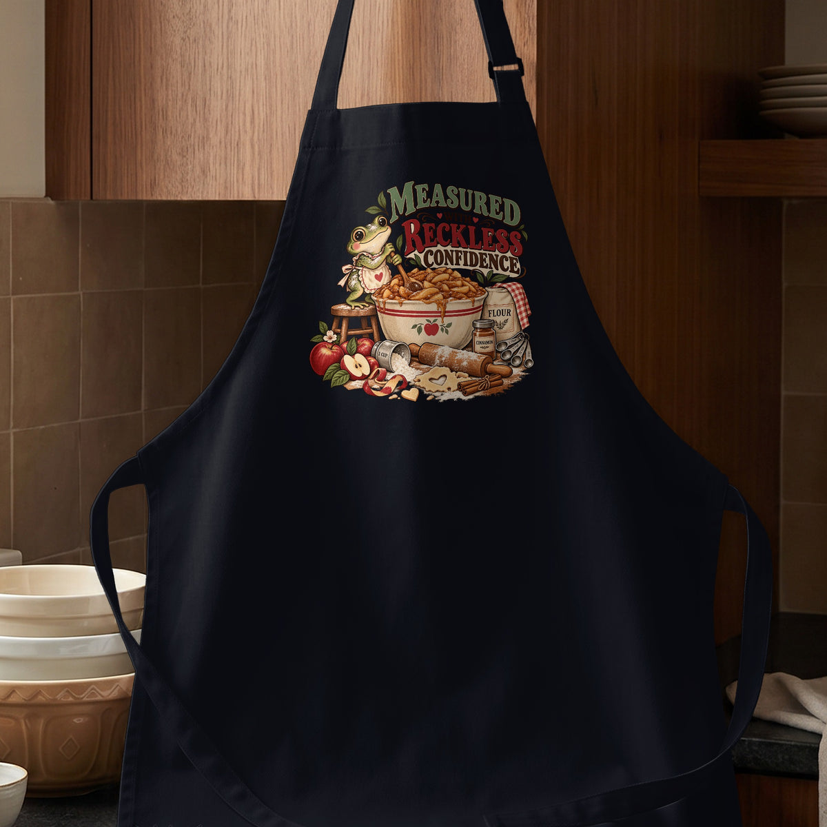 Black apron with a graphic of a frog and baking ingredients, reading "Measured with Reckless Confidence".