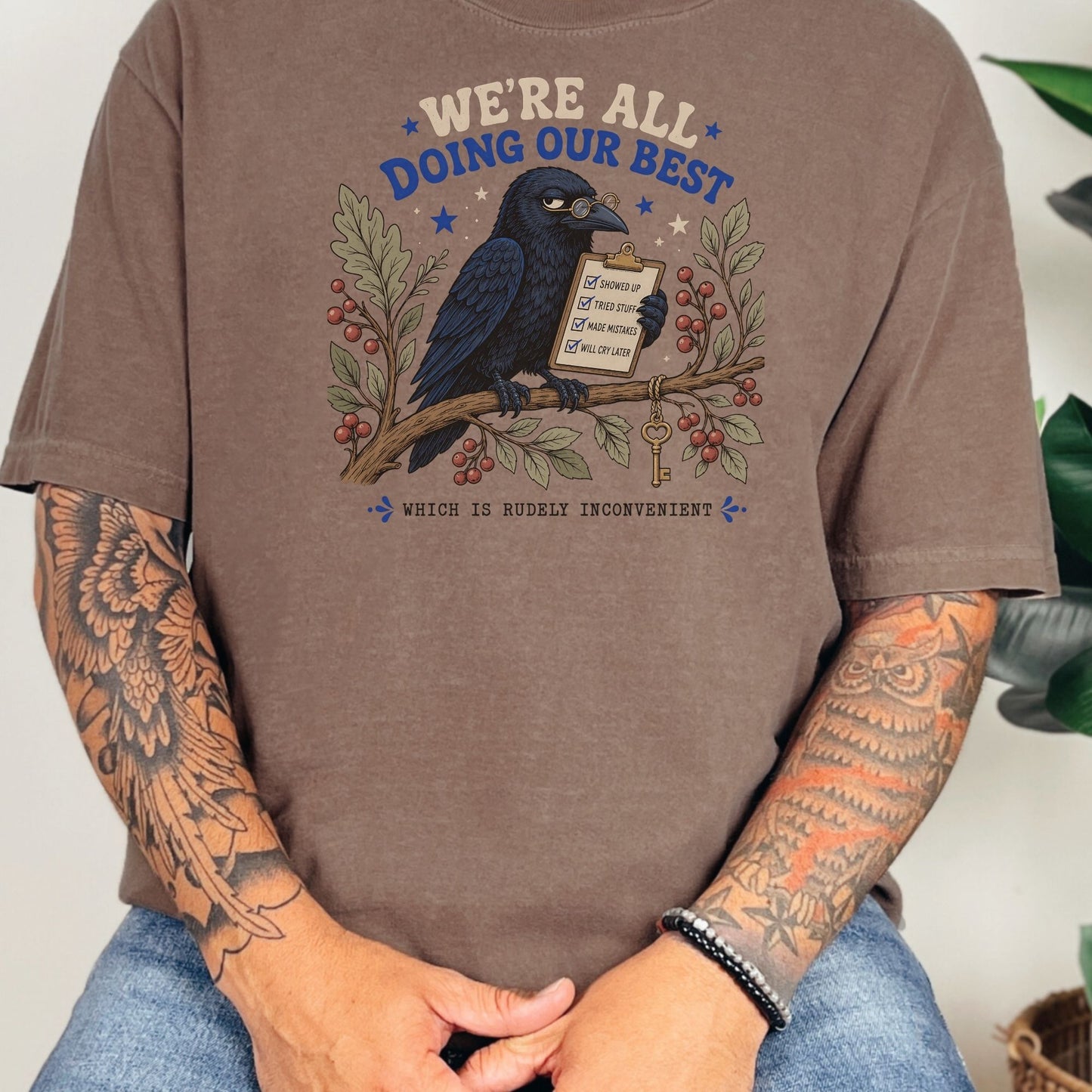 Brown t-shirt with a crow wearing glasses holding a clipboard and branch with berries.