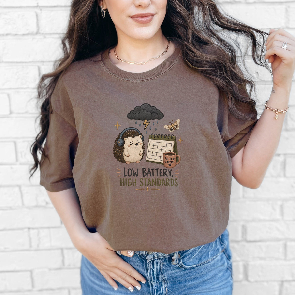 Brown t-shirt with embroidered hedgehog, cloud, calendar, and coffee mug graphic with text.
