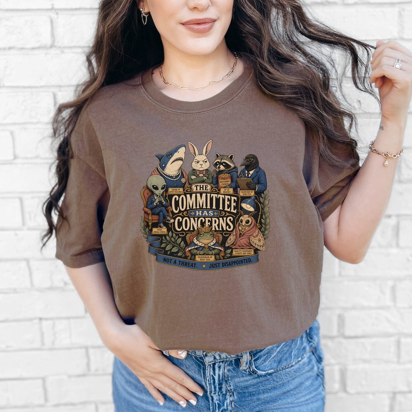 A person wearing a brown crewneck t-shirt featuring a cartoon graphic of animals and an alien surrounding text that reads The Committee Has Concerns.