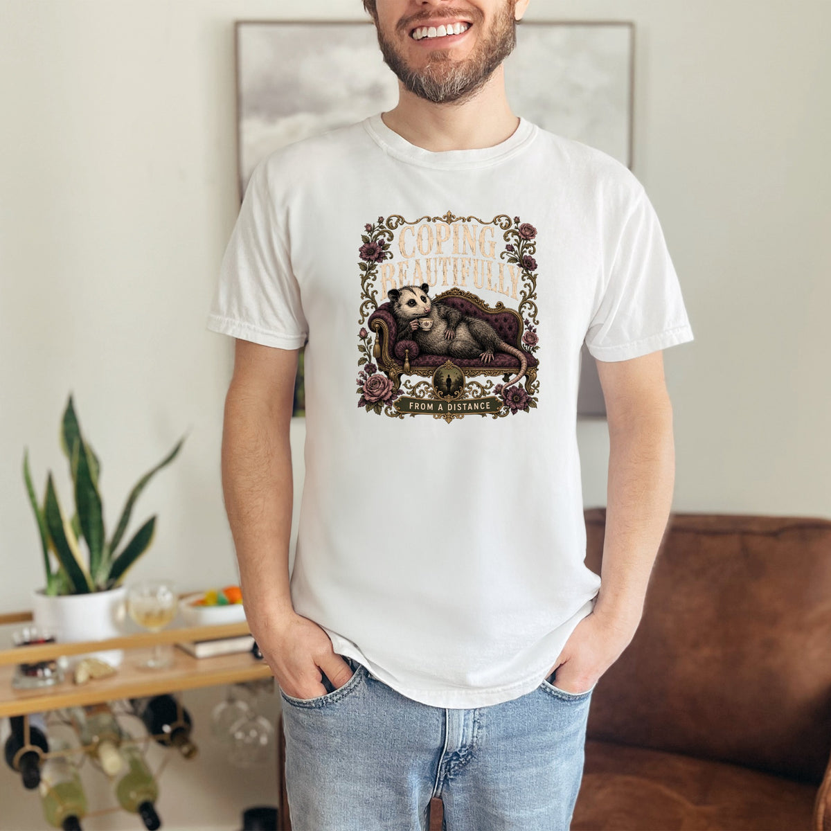White t-shirt with a vintage illustration of an opossum on a couch reading "Coping Beautifully From A Distance".