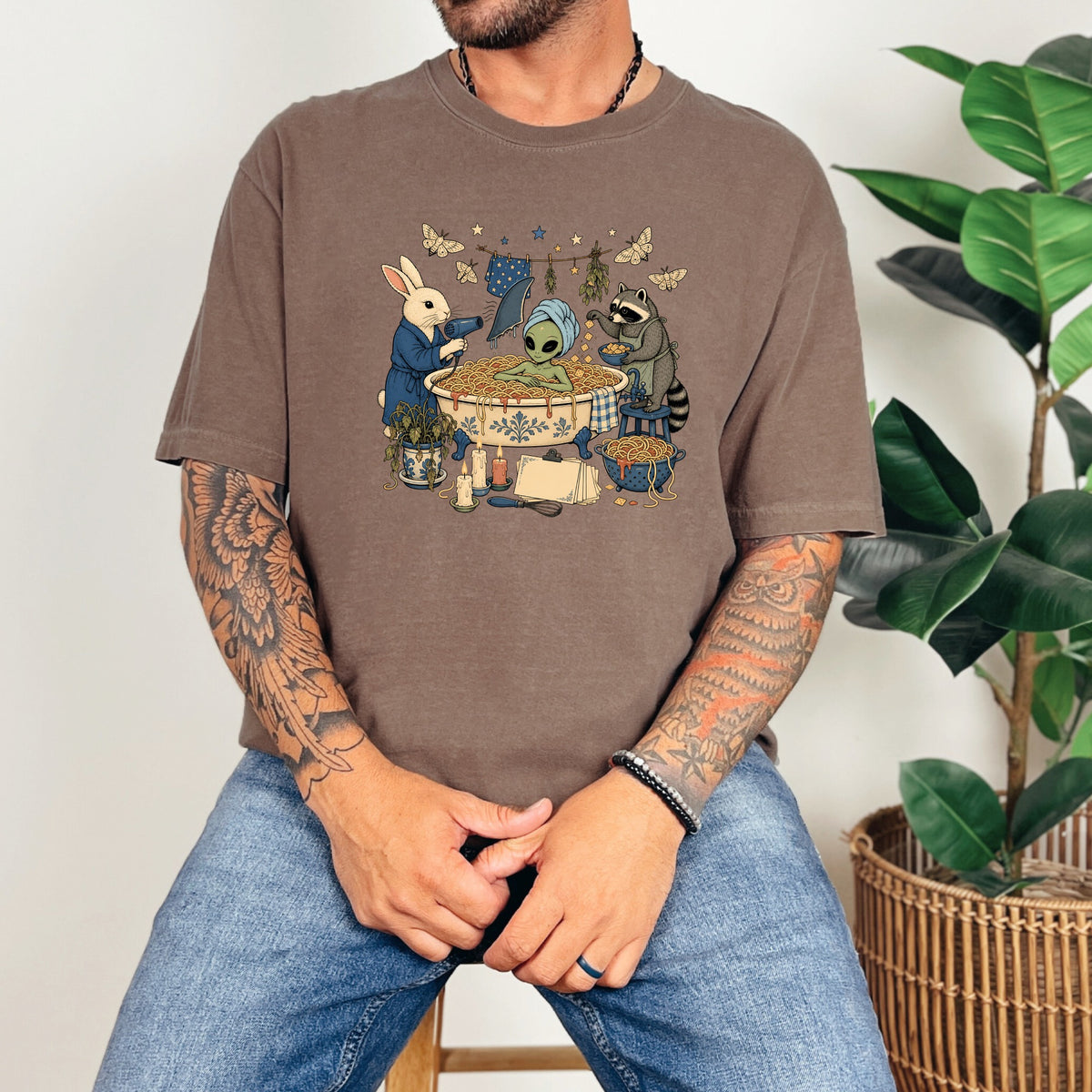 A person wearing a brown t-shirt featuring a graphic of an alien in a spaghetti bathtub with animals.