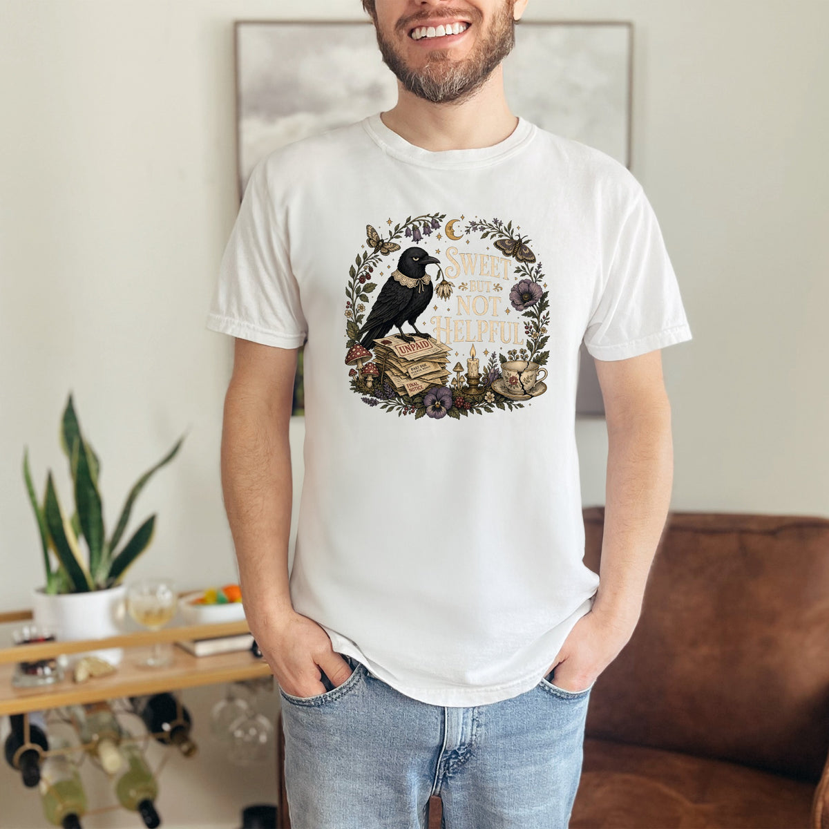 White t-shirt with a circular graphic of a crow, books, and flowers with text "Sweet but Not Helpful".
