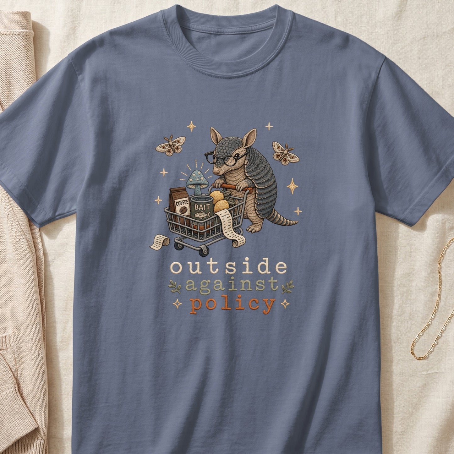 Blue t-shirt with embroidered armadillo pushing a shopping cart with text "outside against policy".