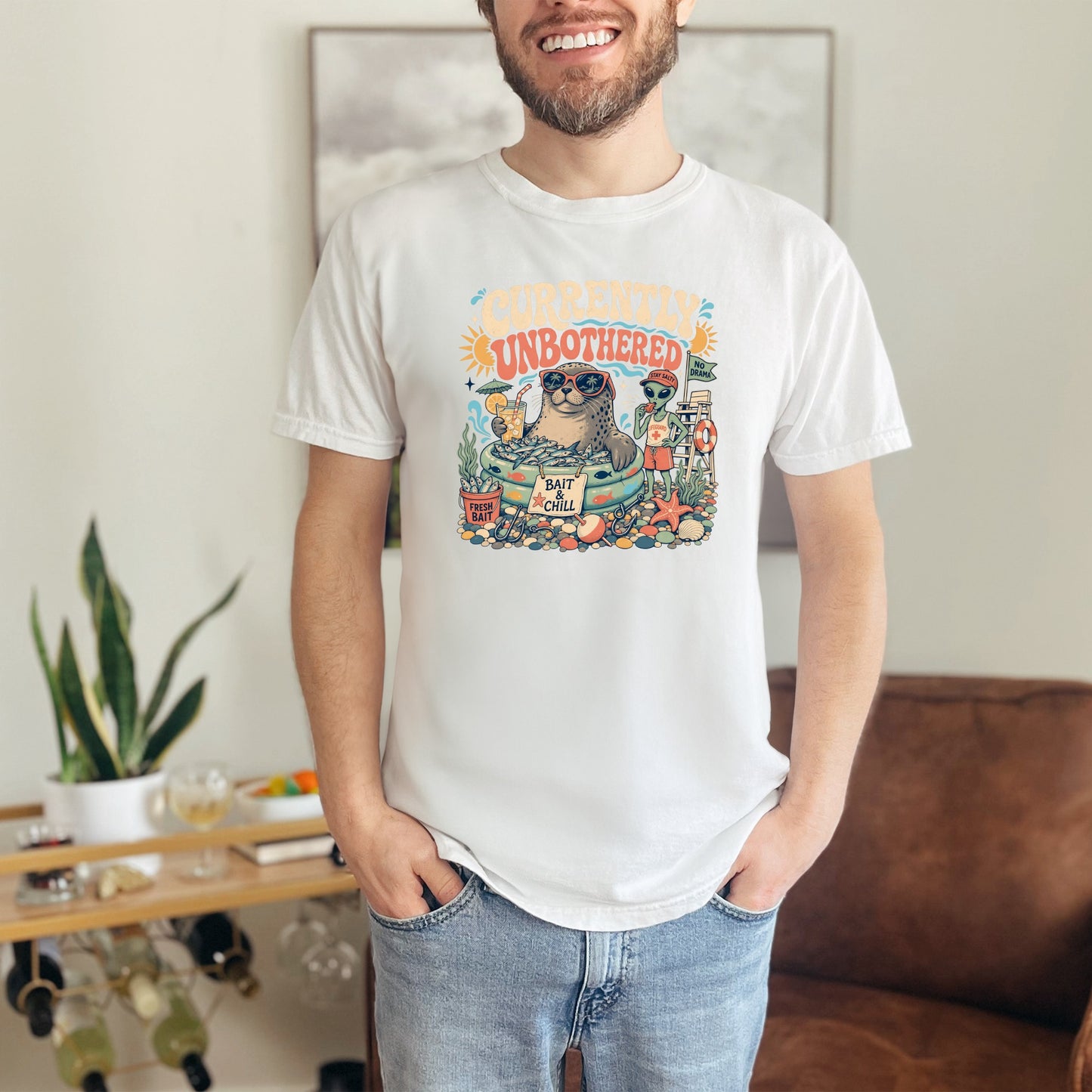 White t-shirt with graphic "Currently Unbothered" featuring a seal, alien, and beach scene.