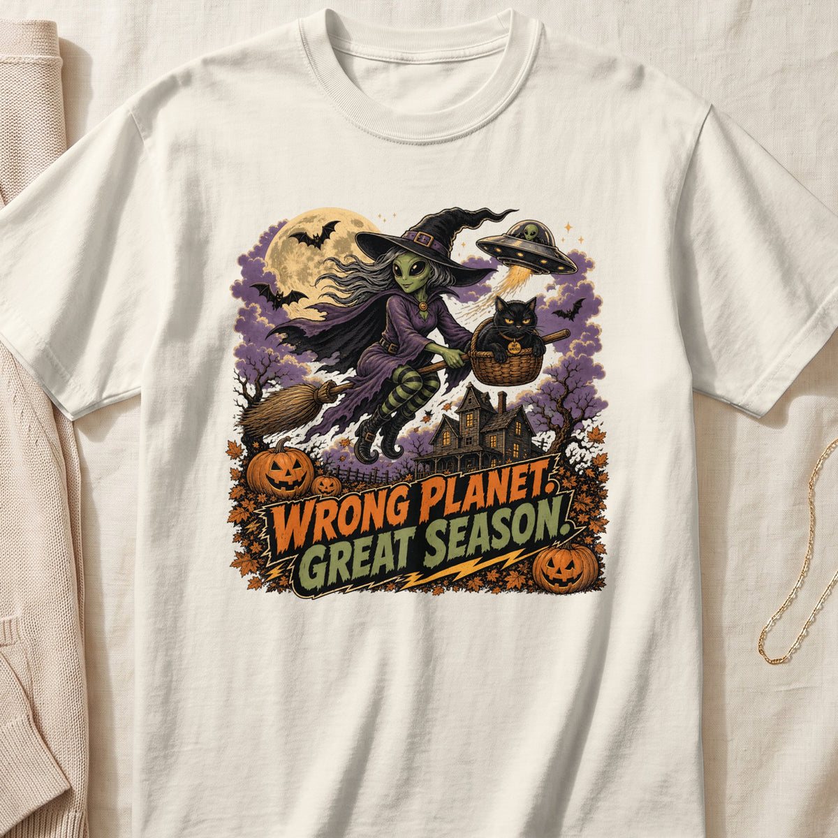 Alien witch with black cat in basket flies past haunted house, text reads "Wrong Planet, Great Season."