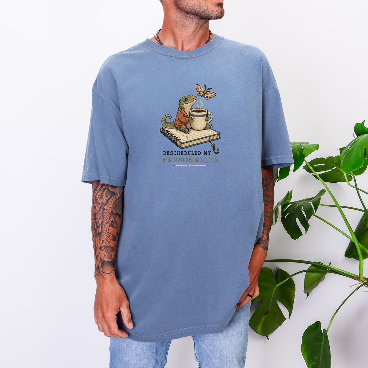 A man wearing an oversized blue t-shirt featuring a lizard drinking coffee on a notebook.