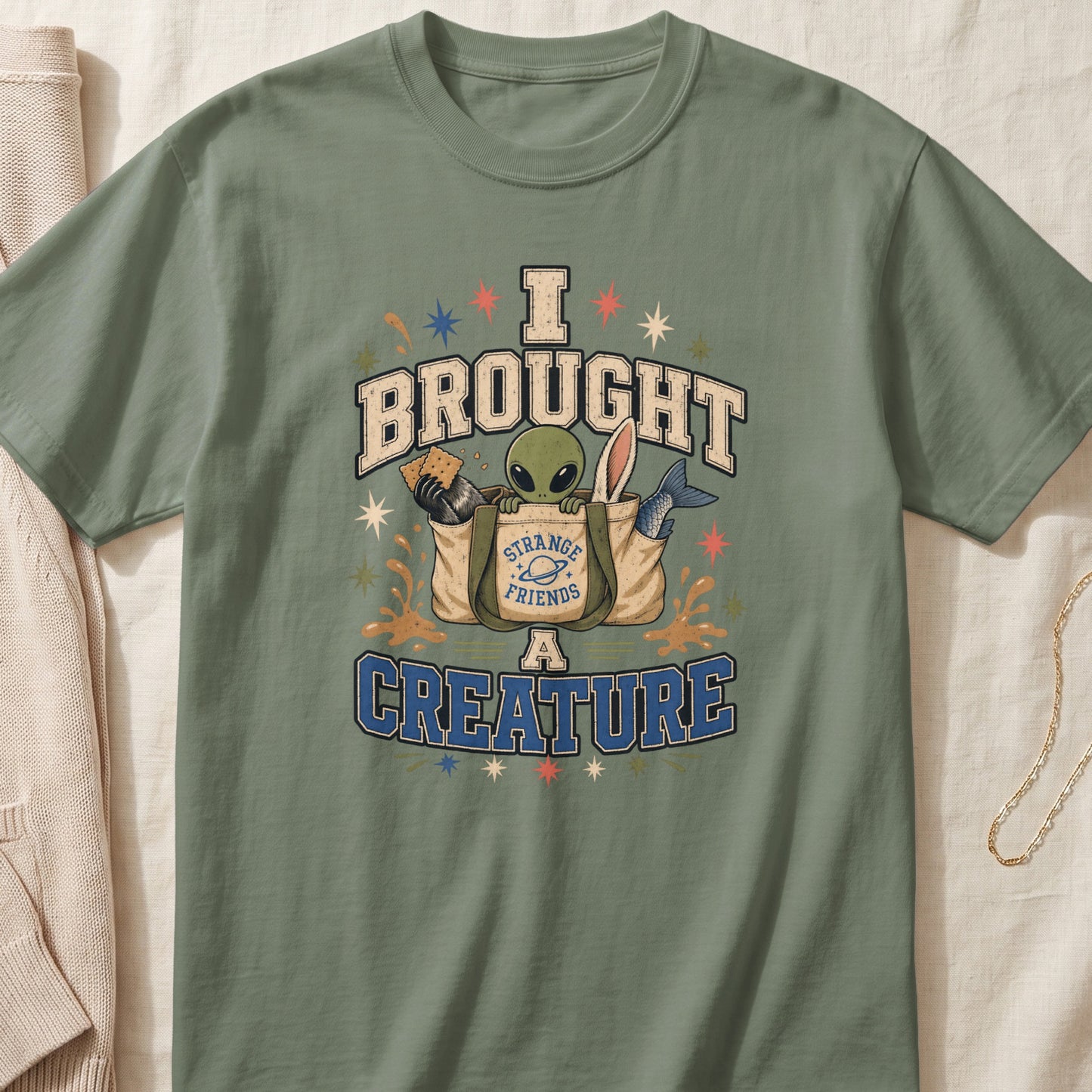 Green t-shirt with text "I BROUGHT A CREATURE" and an alien in a bag of strange friends.