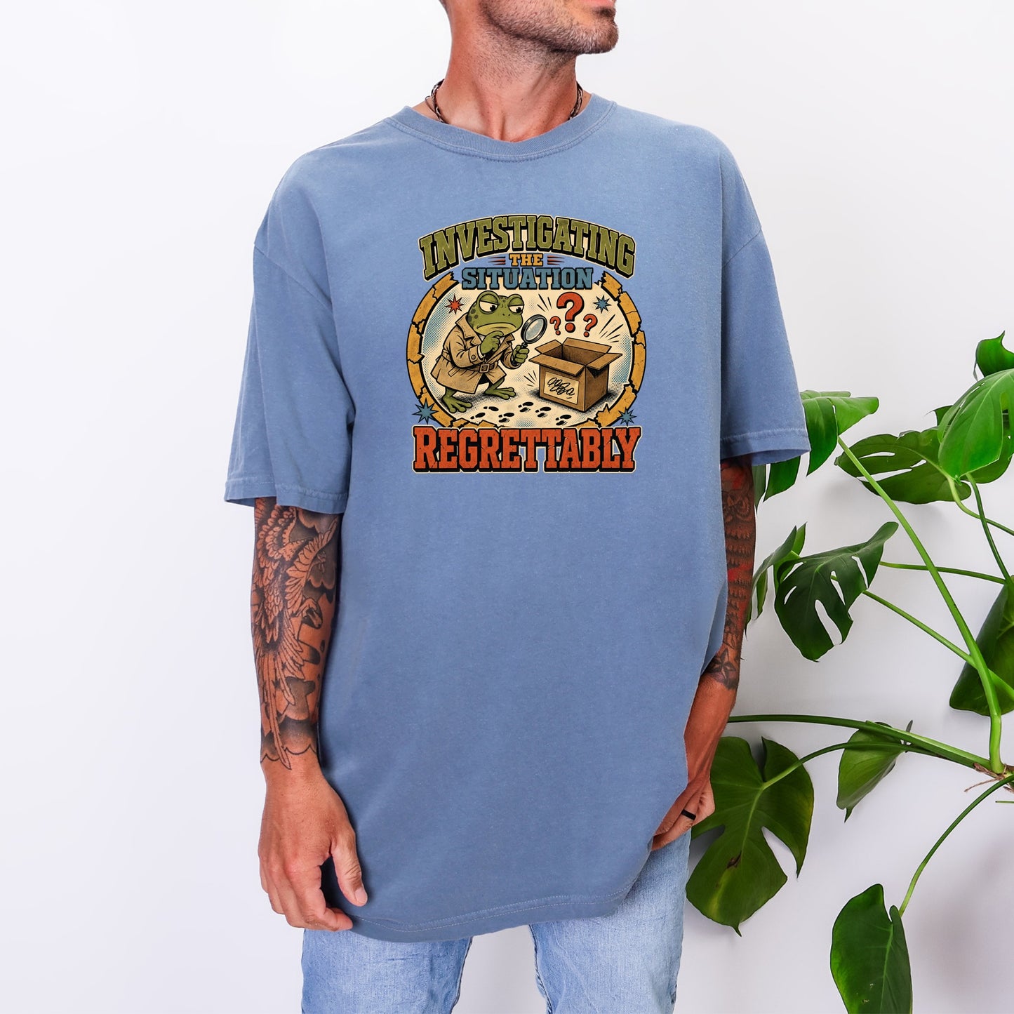 A person wearing an oversized blue t-shirt featuring a graphic of a frog investigating a box with a magnifying glass and the text Investigating the Situation Regrettably.