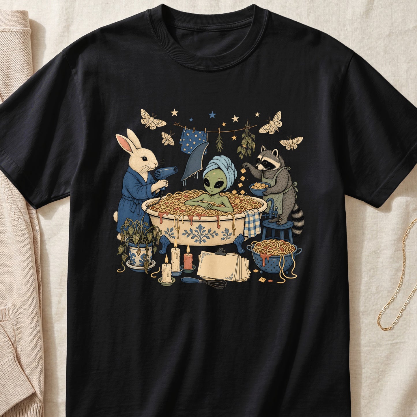 A black t-shirt features a whimsical graphic of an alien in a spaghetti bath tended by a rabbit and raccoon.