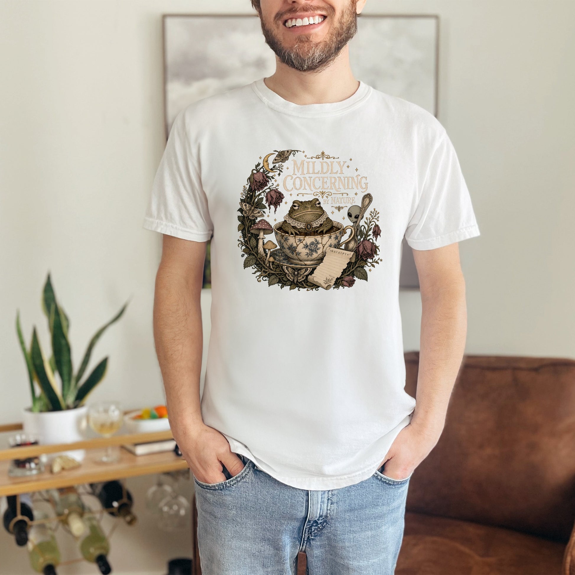 White t-shirt with a circular graphic of a frog in a teacup, mushrooms, and text "Mildly Concerning By Nature".